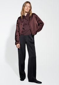 Giacca bomber in satin bordeaux, top in maglia a coste, pantaloni neri a gamba larga e scarpe slip-on nere, su uno sfondo bianco uniforme.