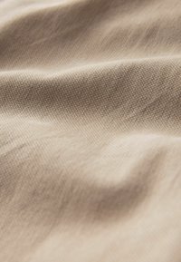 Šviesiai rudas audinys, audžiamas su smulkia tekstūra, rodantis švelnias bangas ir raukšles, pabrėžiantis jo lygų, šiek tiek atspindintį paviršių.