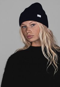 Young woman with long blonde hair, freckles, wearing a black knit beanie and black sweater, looking slightly to the side against gray background.