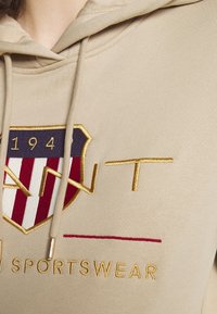 Felpa beige con cappuccio caratterizzata da un logo ricamato con accenti dorati, strisce blu navy e rosse in un design a scudo, e testo ben visibile.