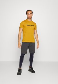 Gul t-shirt med svart "Mammut"-logotyp, kombinerad med grå shorts och marinblå leggings, bärs med svarta träningsskor. Enkelt mönster, figursydd stil.