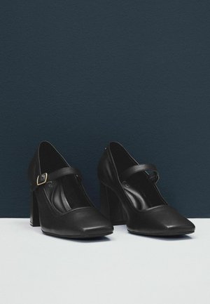 Scarpe nere con tacco, punta quadrata, una larga fascia sul collo del piede e un dettaglio con fibbia dorata. Realizzate in pelle liscia.