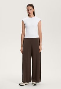 Biele priliehavé tričko s krátkym rukávom v kombinácii s voľnými hnedými širokými nohavicami. Model má obuté šedé a biele športové topánky. Jednoduchý, čistý dizajn.