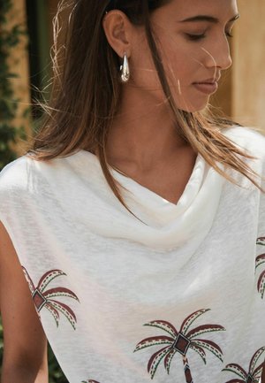 Woman with long brown hair wearing white sleeveless top with palm tree prints and silver teardrop earrings, looking downward to the side.