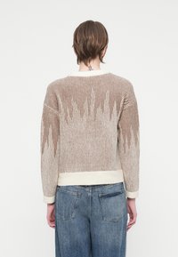 Person med kort brunt hår og nakke-tatovering iført en beige og hvid stribet striktrøje og blå jeans, set bagfra.