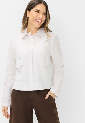 Lächelnde Frau, die eine weiße Jacke mit Knöpfen und zwei Fronttaschen sowie braune Hose trägt und mit einer Hand in der Tasche steht.