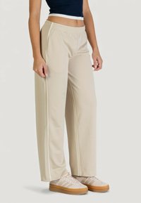 Pantaloni beige a gamba larga realizzati in tessuto liscio, con strisce laterali bianche e vita elastica. Abbinati a sneaker di colore chiaro.