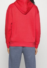 Rote Kapuzenpullover mit langen Ärmeln, lässiger Passform sowie gerippten Bündchen und Saum. Kombiniert mit grauen Hosen mit einem gesprenkelten Muster.