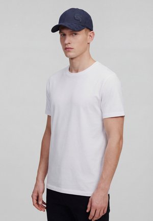 Jonge man die een eenvoudige witte T-shirt met korte mouwen en een marineblauwe baseballpet draagt, staand tegen een effen grijze achtergrond.