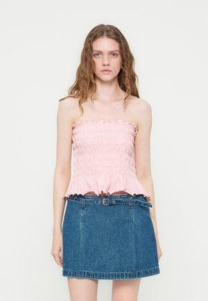 Jonge vrouw met lang golvend haar die een roze strapless gesmokte top en een blauwe denim rok met riem draagt, staand tegen een effen witte achtergrond.