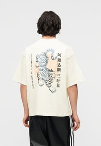 Άτομο με κοντά σκούρα μαλλιά που φοράει μια υπερμεγέθη λευκοκρεμ μπλούζα με γραφικό ενός τίγρη που βρυχάται και κάθετο κείμενο στα κινέζικα και αγγλικά.