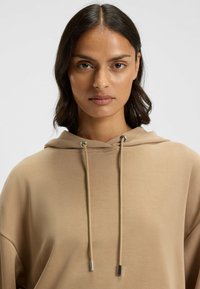 Felpa beige con cappuccio e coulisse, dotata di aghetti metallici. Realizzata in tessuto morbido, con una vestibilità rilassata e senza motivi visibili.