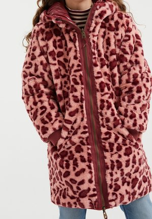 Femme portant un long manteau en fausse fourrure imprimé léopard rose et bordeaux, avec une fermeture éclair devant, les mains dans les poches, et une chemise rayée en dessous.