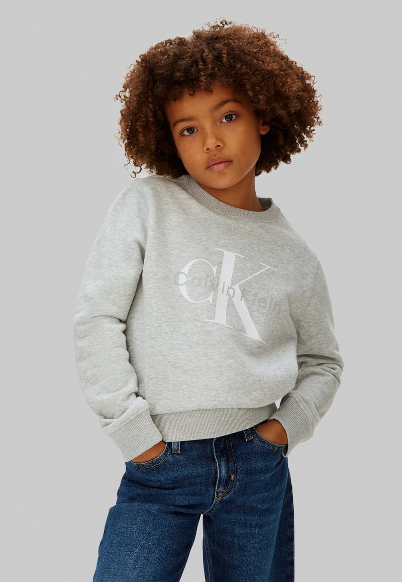 Παιδί με σγουρά μαλλιά που φοράει ανοιχτό γκρι φούτερ Calvin Klein και μπλε τζιν, στέκεται με τα χέρια στις τσέπες μπροστά σε απλό φόντο.