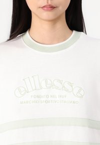 Balts kokvilnas T-krekls ar gaiši zaļu ribotu apkakli un akcentiem. Ar izšūtu "ellesse" logo un tekstu. Mīksta tekstūra.