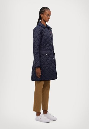 Marineblå quilted frakke med krave, trykknapper, to frontlommer og kontrastkanter, parret med beige bukser og hvide sneakers.
