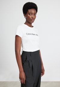 Witte katoenen t-shirt met korte mouwen en een ronde hals, met een zwart "Calvin Klein Jeans" logo op de borst, gecombineerd met high-waisted zwarte broeken.