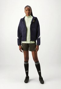Veste zippée marine avec poches avant, couche de base vert clair, shorts olive, chaussettes noires montantes avec des rayures, et chaussures de sport noires.