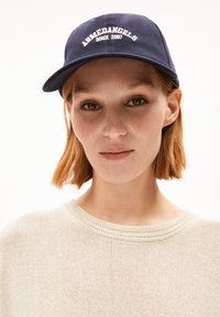 Navy baseball cap with "ARMEDANGELS SINCE 2007" embroidered in white. Soft texture, curved brim, worn with a light beige knit sweater.