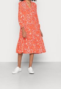 Langarmkleid mit V-Ausschnitt in leuchtendem Orange mit weißen Blumenmustern; ausgestellter Rockschnitt kombiniert mit weißen Turnschuhen. Glatter Stoff.