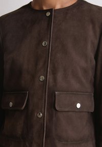 Braune Wildlederjacke mit rundem Halsausschnitt, vorderen Druckknopfverschlüssen und zwei Brusttaschen mit silbernen Akzenten und Stickdetails.