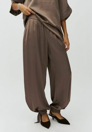 Person trägt lockere braune Satin-Hose mit elastischen Bündchen, die an den Knöcheln gebunden sind, und passendes übergroßes Oberteil, kombiniert mit dunklen spitzen Slingback-Schuhen.