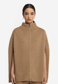 Kamelfärgad stickad poncho med hög krage, dragkedja och öppna sidor. Ribbad fåll ger textur och detalj åt designen.