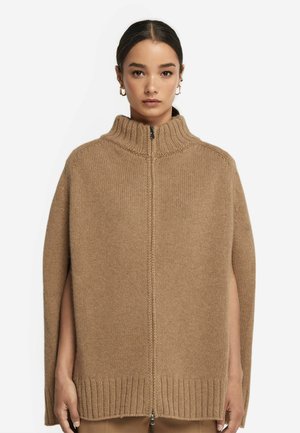 Kamelfärgad stickad poncho med hög krage, dragkedja och öppna sidor. Ribbad fåll ger textur och detalj åt designen.