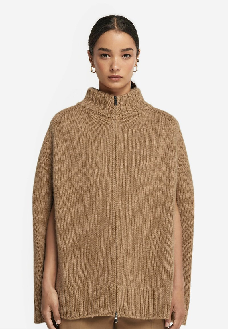 Kamelfärgad stickad poncho med hög krage, dragkedja och öppna sidor. Ribbad fåll ger textur och detalj åt designen.