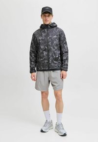En ung mand står iført en sort og grå mønstret hættetrøje, lysegrå shorts, hvide sportssokker, grå sneakers og en sort cap.