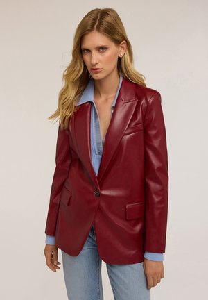 Blazer en cuir rouge avec un col cranté, fermeture à un bouton et deux poches avant. Porté sur une chemise texturée bleu clair et un jean.