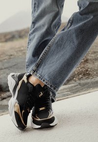 Svarta och gyllene träningsskor i mesh och mocka, med en kraftig vit sula och metalliska detaljer. Bärs med blå jeans.