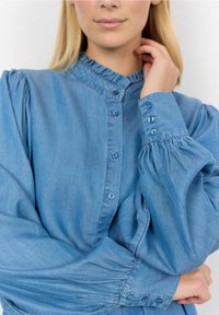 Camicia di jeans azzurro chiaro con colletto arricciato, chiusura frontale con bottoni e maniche a sbuffo con dettagli di bottoni sui polsini. Texture morbida.