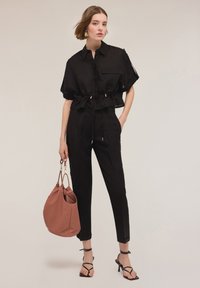 Cropped nero con vita a coulisse, abbinato a pantaloni neri su misura. La modella tiene una borsa marrone testurizzata e indossa tacchi neri con cinturini.