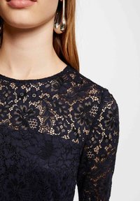 Haut en dentelle marine avec motif floral, doté d'un col rond et de manches longues. Superposition en dentelle texturée et tissu uni à la base.
