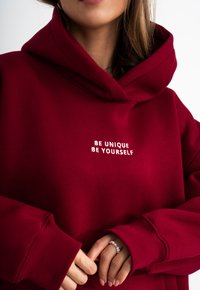 Felpa bordeaux con un grande cappuccio piegato. Presenta del testo ricamato bianco "ESSERE UNICO ESSERE SE STESSO" sul petto. Tessuto morbido, vestibilità comoda.