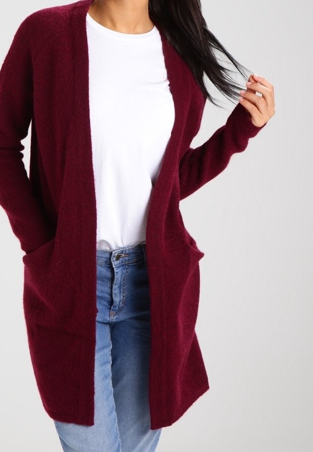 Femme portant un long cardigan bordeaux par-dessus un t-shirt blanc et un jean bleu, une main dans la poche et l'autre touchant ses cheveux.