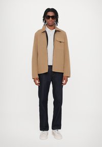 Beige Jacke mit Reißverschluss vorne, einer Brusttasche, kombiniert mit einem hellgrauen Poloshirt, dunkelblauen Jeans und weißen Sneakers.