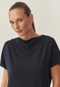 Marineblaues, locker sitzendes Oberteil mit einem drapierten Ausschnitt und kurzen Ärmeln, aus weichem Stoff, der eine glatte Textur bietet. Goldene Ohrringe ergänzen den Look.