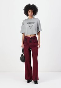 Maglietta corta grigia con logo triangolo grafico nero. Jeans a gamba larga rosso scuro con tasche frontali. Modello porta una piccola borsa nera.
