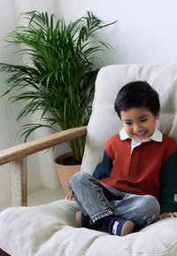 Kind sitzt auf einem hellbeigen, gepolsterten Stuhl, trägt ein rot-navyfarbenes Langarm-Poloshirt und blaue Jeans, im Hintergrund eine grüne Pflanze.