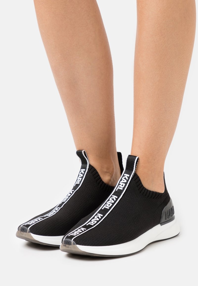 Pieds portant des baskets noires en maille slip-on avec le texte blanc « KARL » sur les languettes verticales, dotées de semelles blanches et de bouts translucides.