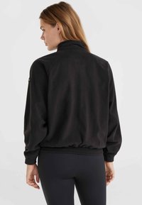 Veste en polaire noire avec un col haut, des poignets élastiques et une taille élastique ; elle présente un design à épaule tombante et un petit logo sur la manche gauche.