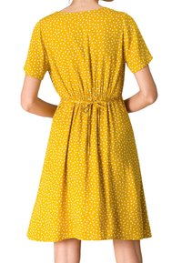 Femme portant une robe jaune à manches courtes avec des pois blancs, nouée à la taille, vue de dos.