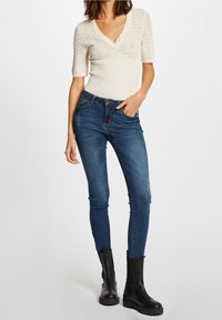 Krämfärgad ribbad stickad topp med korta ärmar och V-ringning, parat med mörkblå skinny jeans med lätt slitage och svarta ankelboots.