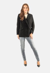 Donna con lunghi capelli castani che indossa un blazer nero, un maglione chiaro, jeans grigi e tacchi neri, in piedi con una gamba piegata.