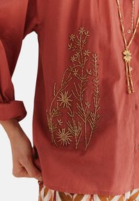 Rustfarbenes Hemd mit Knopfleiste, das auf der Vorderseite mit kunstvollen goldenen Stickereien von floralen Designs verziert ist, mit hochgerollten Ärmeln und einem lässigen Schnitt.