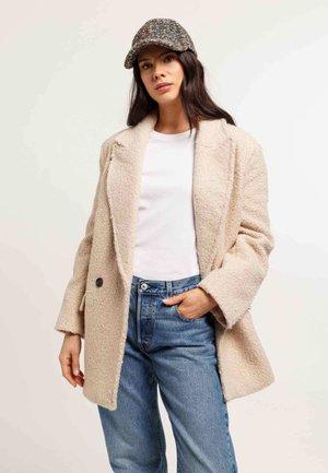 Cappotto oversize beige testurizzato con ampi rever e bottoni neri, indossato su una camicia bianca e jeans in denim blu, abbinato a un berretto a fantasia.