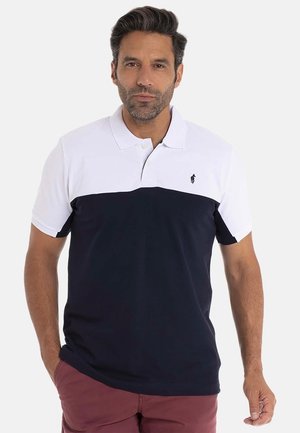 Homme portant un polo blanc et marine à manches courtes avec un petit logo brodé, associé à un pantalon bordeaux, se tenant devant un fond uni.