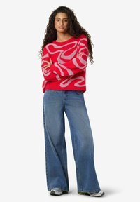 Rdeč pulover z rožnatim valovitim vzorcem, sproščen kroj in dolgi rokavi, kombiniran z modrimi kavbojkami širokih nog z ozkim pasom in sivimi supergami.